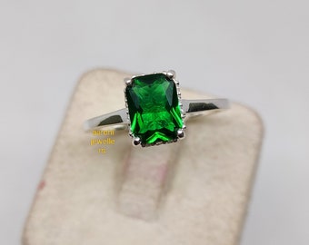 Puede incluir: Un anillo de plata con una gran piedra preciosa verde esmeralda. La piedra preciosa está engastada en un engaste cuadrado de garras.