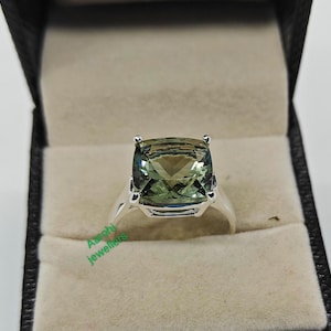 Puede incluir: Un anillo de plata con una gran piedra preciosa verde de forma cuadrada. El anillo está en una caja de anillo negra y blanca.