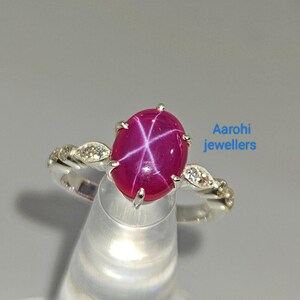 Pode incluir: Um anel de prata com uma gema de rubi estrela rosa e pequenos diamantes engastados na banda. O anel está em um expositor branco. Aarohi jewellers.
