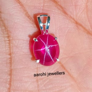 Pode incluir: Um pingente de rubi estrela vermelho com uma configuração de prata. O rubi é oval e tem um padrão de estrela no centro.