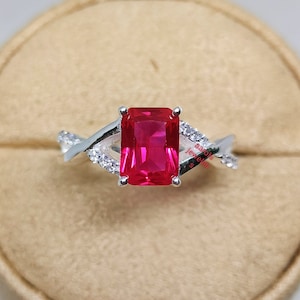 May include: A silver ring with a large, red, emerald-cut gemstone surrounded by smaller, clear gemstones. The ring is on a beige display stand.