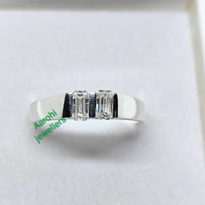 Puede incluir: Un anillo de plata con dos piedras preciosas rectangulares y transparentes, colocadas una al lado de la otra. El anillo tiene una banda lisa y pulida. El anillo se muestra sobre una superficie blanca.