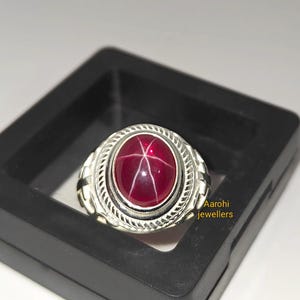 May include: A silver ring with a large, oval, deep red gemstone. The gemstone has a star-like pattern across its surface. The ring is set in a decorative silver band and is displayed in a black box.