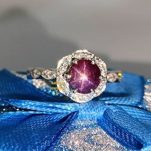 May include: A silver ring with a large, round, red star ruby gemstone surrounded by smaller white diamonds. The ring is set on a blue velvet box.