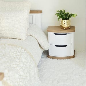 May include: A white two-drawer bedside table with a light brown top and base, featuring curved drawer fronts with black recessed handles. A small green plant in a textured gold pot sits on the surface next to a bed with a cream-coloured textured quilt.