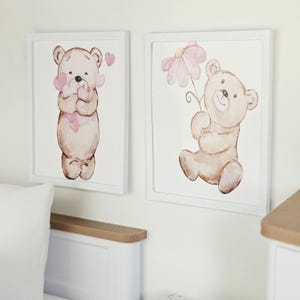 May include: Two square framed wall art pieces featuring watercolour illustrations of light brown teddy bears. One bear holds a cluster of pink hearts, while the other bear holds a single large pink flower. Both prints are set in white frames.