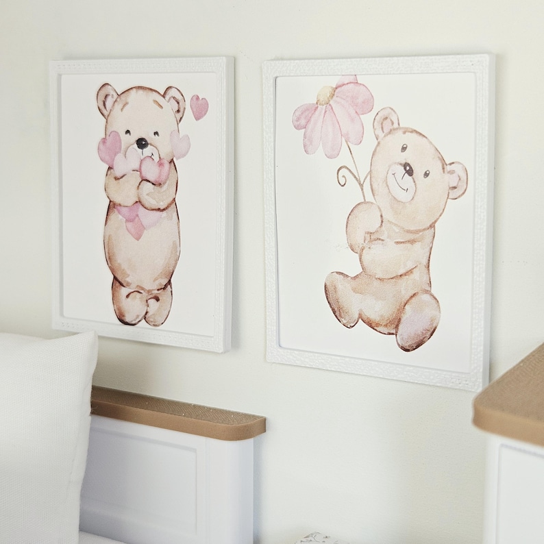 May include: Two square framed wall art pieces featuring watercolour illustrations of light brown teddy bears. One bear holds a cluster of pink hearts, while the other bear holds a single large pink flower. Both prints are set in white frames.