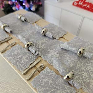 May include: A set of five place settings with silver napkin rings and gray patterned linen napkins. The place settings are arranged on a wooden table.