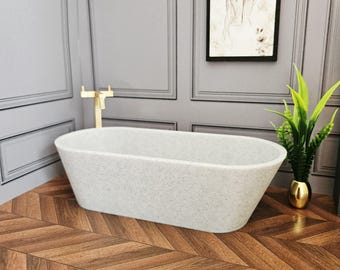 May include: A miniature, speckled gray bathtub with an oval shape. The tub is set in a bathroom scene with a gold faucet, a framed picture, and a small potted plant. The floor has a herringbone pattern.