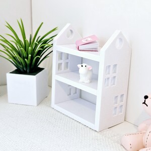 May include: White wooden shelf unit shaped like a house with two shelves. The top shelf has a pink book on it and the bottom shelf has a small white lamb figurine.