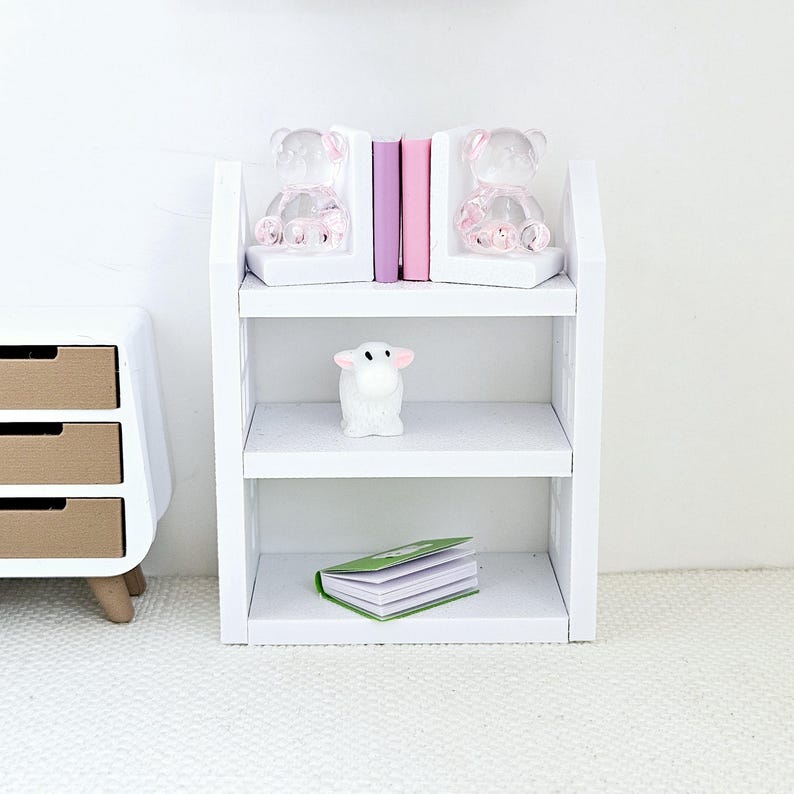 May include: White bookshelf with two shelves. The top shelf has two pink teddy bear bookends and three books. The middle shelf has a white lamb figurine. The bottom shelf has a green and white book.