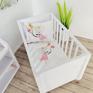 May include: A white doll-sized crib with a matching pillow and blanket set. The bedding features a cartoon mouse wearing a crown and pink dress, holding a star and bird. The crib is made of wood and has a slatted side.