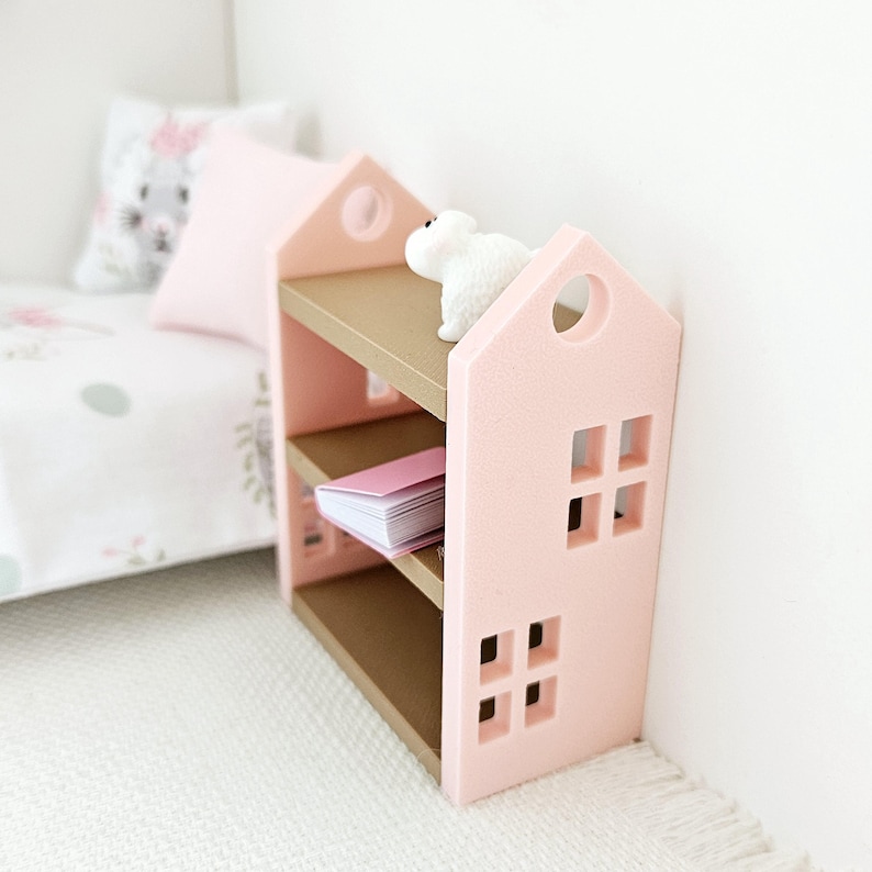 May include: A pink wooden bookshelf with two shelves and a white animal figurine on top. The bookshelf has a house-shaped design with cut-out windows.