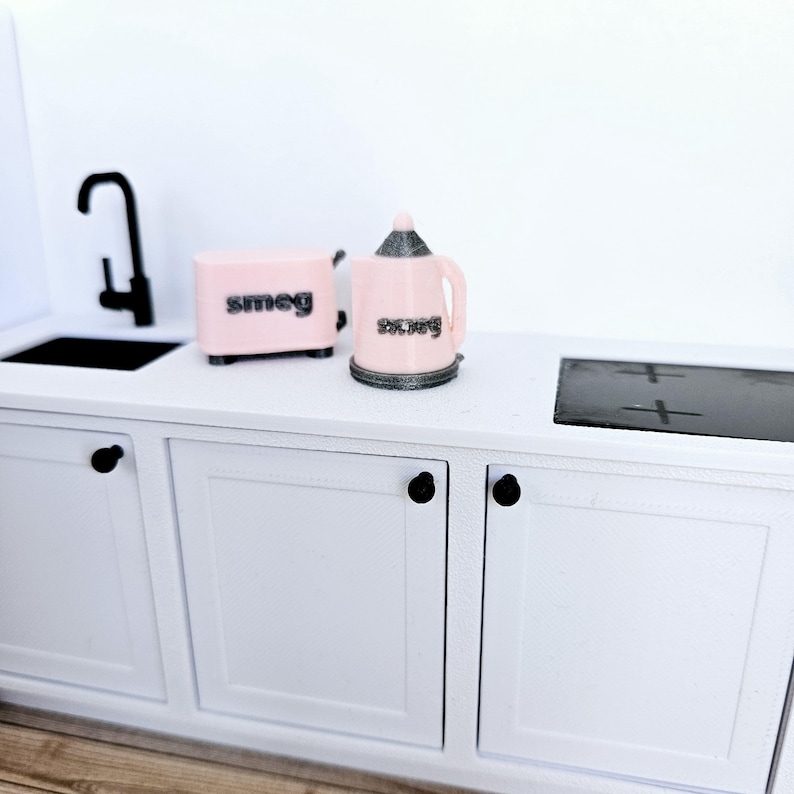May include: A white kitchen cabinet with two doors and black knobs. A pink toaster and a pink kettle with the word "smeg" on them are on the counter.