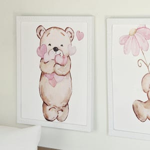 May include: A rectangular wall art piece in a white frame featuring a watercolour illustration of a light brown teddy bear holding a cluster of soft pink hearts. The bear has a simple, rounded design with a small black nose and dark eyes.