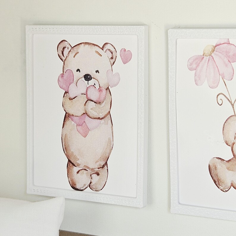 May include: A rectangular wall art piece in a white frame featuring a watercolour illustration of a light brown teddy bear holding a cluster of soft pink hearts. The bear has a simple, rounded design with a small black nose and dark eyes.