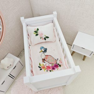 Pode incluir: Um berço branco com um conjunto de roupa de cama rosa e floral com um pato com um gorro azul. O conjunto de roupa de cama inclui um travesseiro e um cobertor com um design floral.