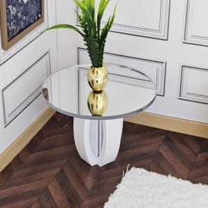 Pode incluir: Uma mesa lateral moderna com tampo espelhado e base branca. Apresenta um vaso dourado com folhas verdes. A mesa está sobre um chão de madeira escura, com painéis brancos no fundo.