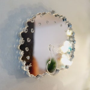 May include: A silver-colored, round mirror with a decorative, scalloped edge. The mirror reflects a blurry image of a room with lights. A peacock design is etched into the mirror.