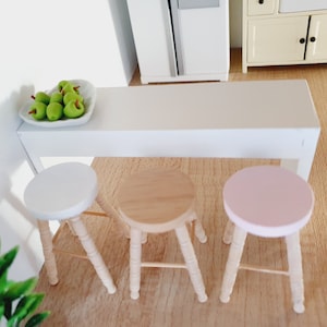 May include: A white table with three wooden stools. The stools have different coloured seats: white, natural wood, and pink. A white bowl with green apples sits on the table.
