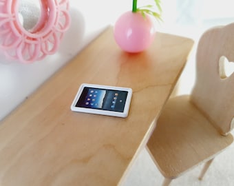Puede incluir: Una tableta blanca con una pantalla negra que muestra un fondo azul y blanco. La tableta está sobre una mesa de madera marrón claro.