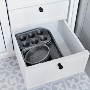 Pode incluir: Uma caneca de cerâmica branca com uma alça preta e o texto "But First, Coffee" em negrito preto. A caneca está sobre um fundo desfocado, sugerindo um ambiente acolhedor.