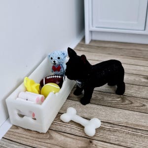May include: A white toy bin with a handle, filled with a variety of toys, including a teddy bear, a football, and a yellow ball. A black French bulldog toy is standing next to the bin, looking at the toys inside. A white bone toy is on the floor.