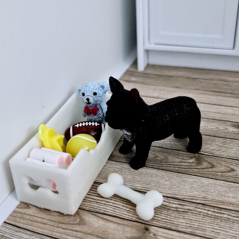 May include: A white toy bin with a handle, filled with a variety of toys, including a teddy bear, a football, and a yellow ball. A black French bulldog toy is standing next to the bin, looking at the toys inside. A white bone toy is on the floor.