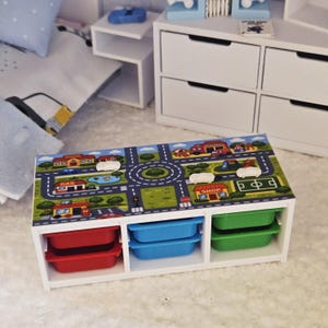 May include: A white storage unit with three compartments, each holding a colorful bin. The top of the unit features a road map design with cars, buildings, and a roundabout. The bins are red, blue, and green.