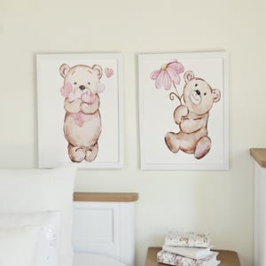 May include: Two rectangular white-framed wall art prints featuring watercolour illustrations of light brown teddy bears. The left print shows a bear holding pink hearts, while the right print depicts a bear holding a large pink flower blossom.
