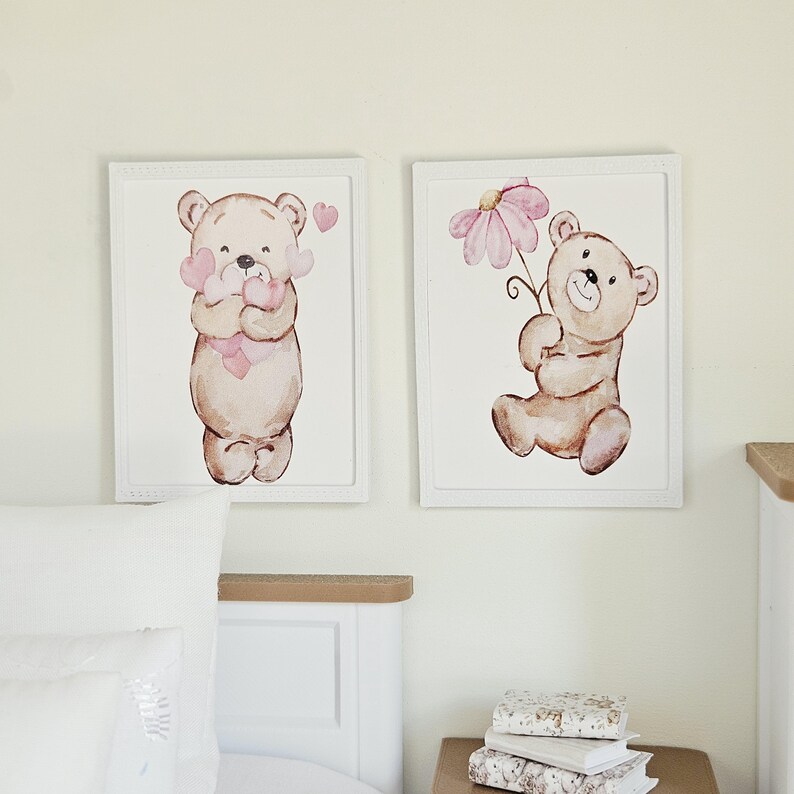 May include: Two rectangular white-framed wall art prints featuring watercolour illustrations of light brown teddy bears. The left print shows a bear holding pink hearts, while the right print depicts a bear holding a large pink flower blossom.