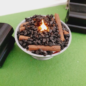 K&ouml;nnte beinhalten: Ein kleiner Tischfeuerplatz mit einer k&uuml;nstlichen Flamme, schwarzen Lavasteinen und braunen Holzscheiten. Der Feuerplatz steht auf einer gr&uuml;nen Oberfl&auml;che.