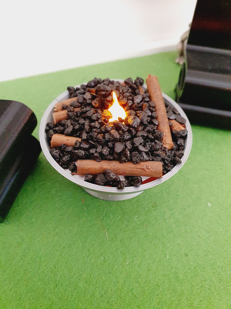 K&ouml;nnte beinhalten: Ein kleiner Tischfeuerplatz mit einer k&uuml;nstlichen Flamme, schwarzen Lavasteinen und braunen Holzscheiten. Der Feuerplatz steht auf einer gr&uuml;nen Oberfl&auml;che.