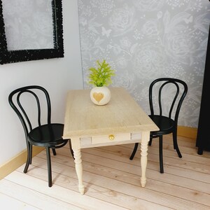 Könnte beinhalten: Ein Miniatur-Esszimmer-Set mit einem hellen Holztisch, einer Schublade und einem goldenen Knopf, flankiert von zwei schwarzen Stühlen. Eine kleine Vase mit Herzmuster und grüner Pflanze steht auf dem Tisch. Die Szene spielt in einem Puppenhaus.