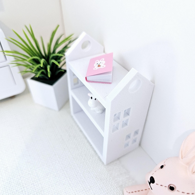 May include: White miniature bookshelf with two shelves, a pink book with a bunny on the cover, and a white figurine on the bottom shelf.