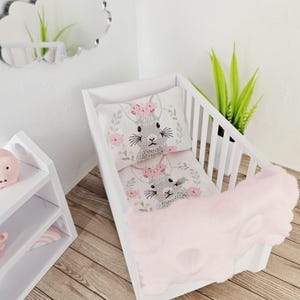 May include: A white doll crib with a pink blanket and a pillow and sheet set featuring a gray bunny with a floral crown. The crib is next to a white shelf and a cloud-shaped mirror.