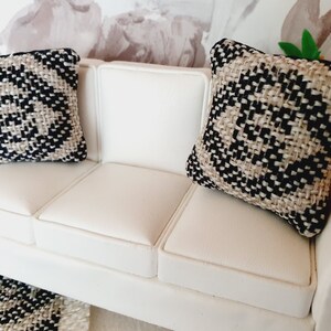 May include: Miniature white sofa with two decorative pillows. The pillows have a woven pattern in black and beige. The sofa is made of white leather-like material. The pillows are square-shaped.