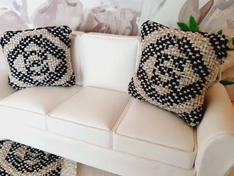 May include: Miniature white sofa with two decorative pillows. The pillows have a woven pattern in black and beige. The sofa is made of white leather-like material. The pillows are square-shaped.