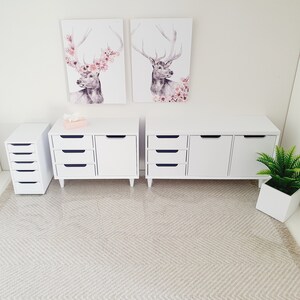 Puede incluir: Dos c&oacute;modas blancas con cajones y puertas, una c&oacute;moda blanca peque&ntilde;a con cajones y una alfombra blanca con un patr&oacute;n de espiga. Dos impresiones enmarcadas de ciervos con acentos florales cuelgan en la pared sobre las c&oacute;modas.