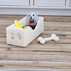 May include: White wooden toy dog bed with a bone cutout on the front. The bed is filled with a blue teddy bear, a yellow toy, and a football.