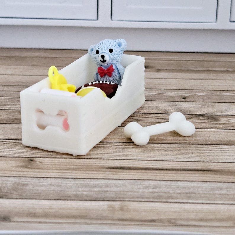 May include: White wooden toy dog bed with a bone cutout on the front. The bed is filled with a blue teddy bear, a yellow toy, and a football.