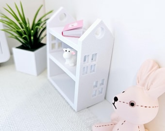 May include: White miniature bookshelf with two shelves, a pink book, a white lamb figurine, and a white background.