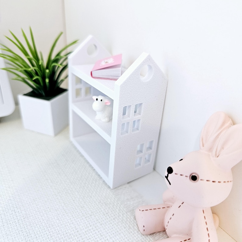 May include: White miniature bookshelf with two shelves, a pink book, a white lamb figurine, and a white background.