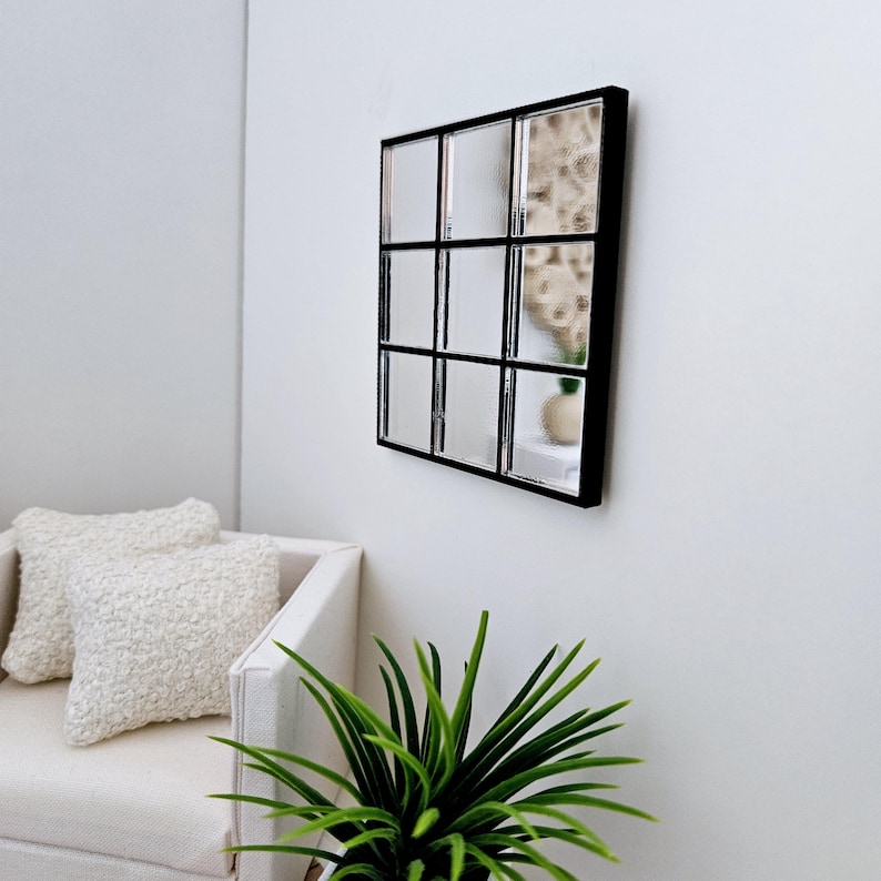 May include: A black framed mirror with nine square panes of glass. The mirror is hanging on a white wall above a white sofa with two white cushions. A green plant is in the foreground.