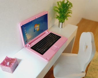 Puede incluir: Un ordenador portátil rosa con un teclado negro y una pantalla blanca que muestra el logotipo de Windows está sobre un escritorio blanco. Una pequeña planta en maceta y una vela rosa también están sobre el escritorio. Una silla blanca está en el fondo.