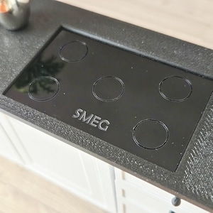 Peut inclure: Une plaque de cuisson SMEG noire avec cinq brûleurs circulaires est encastrée dans un plan de travail gris foncé. La plaque de cuisson est entourée d'une surface texturée, et le logo SMEG est visible. La plaque de cuisson est installée au-dessus de meubles blancs.