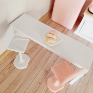May include: A white table with two white chairs and a plate of pink and white eclairs on a white plate.