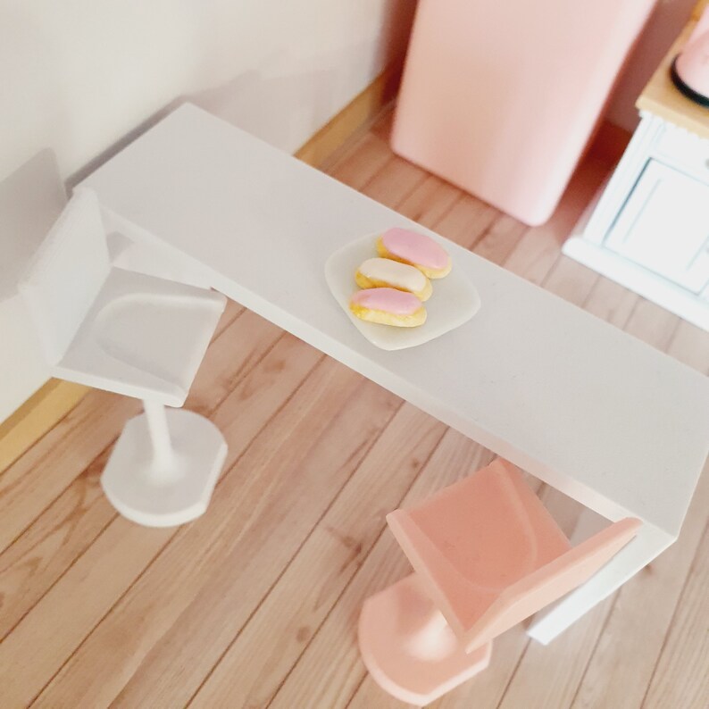 May include: A white table with two white chairs and a plate of pink and white eclairs on a white plate.