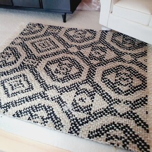 May include: A small, woven rug with a geometric pattern in black and beige. The rug has a fringed edge and is placed on a light-colored surface. The design features repeating diamond and circular shapes.