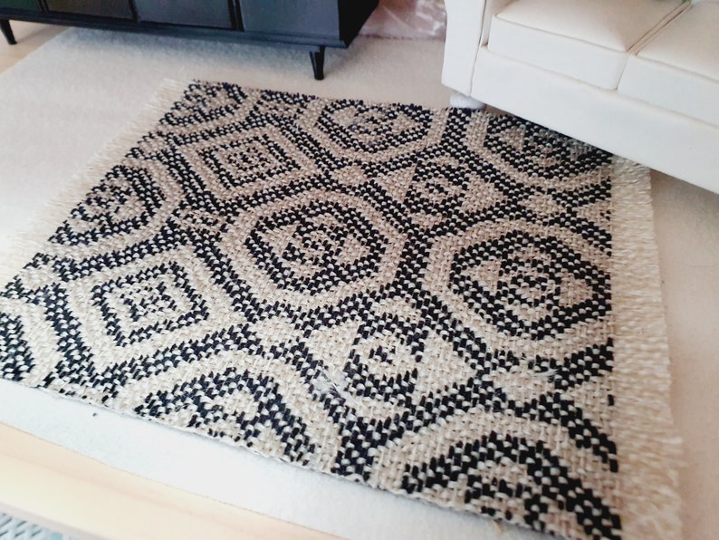 May include: A small, woven rug with a geometric pattern in black and beige. The rug has a fringed edge and is placed on a light-colored surface. The design features repeating diamond and circular shapes.